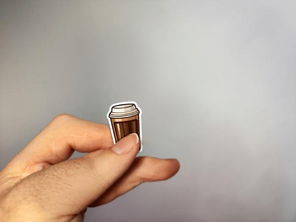 Hand, die einen Aufkleber in Form eines braunen To-Go-Kaffeebechers hält, gestaltet von DreamArt VM.